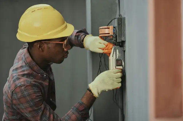 Stock photograph: a tradesperson in a hard hat working at a wall-mounted electrical box. Not a dispatched contractor.